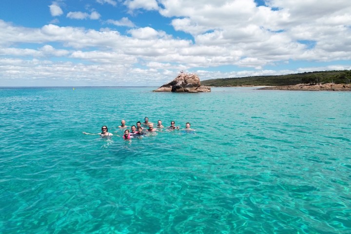 Group swimming in clear blue sea near rocky shore under cloudy sky.