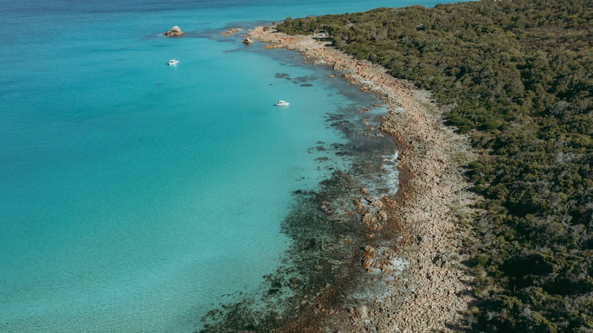 Aerial view of a rocky coastline with turquoise water and two boats near the shore.