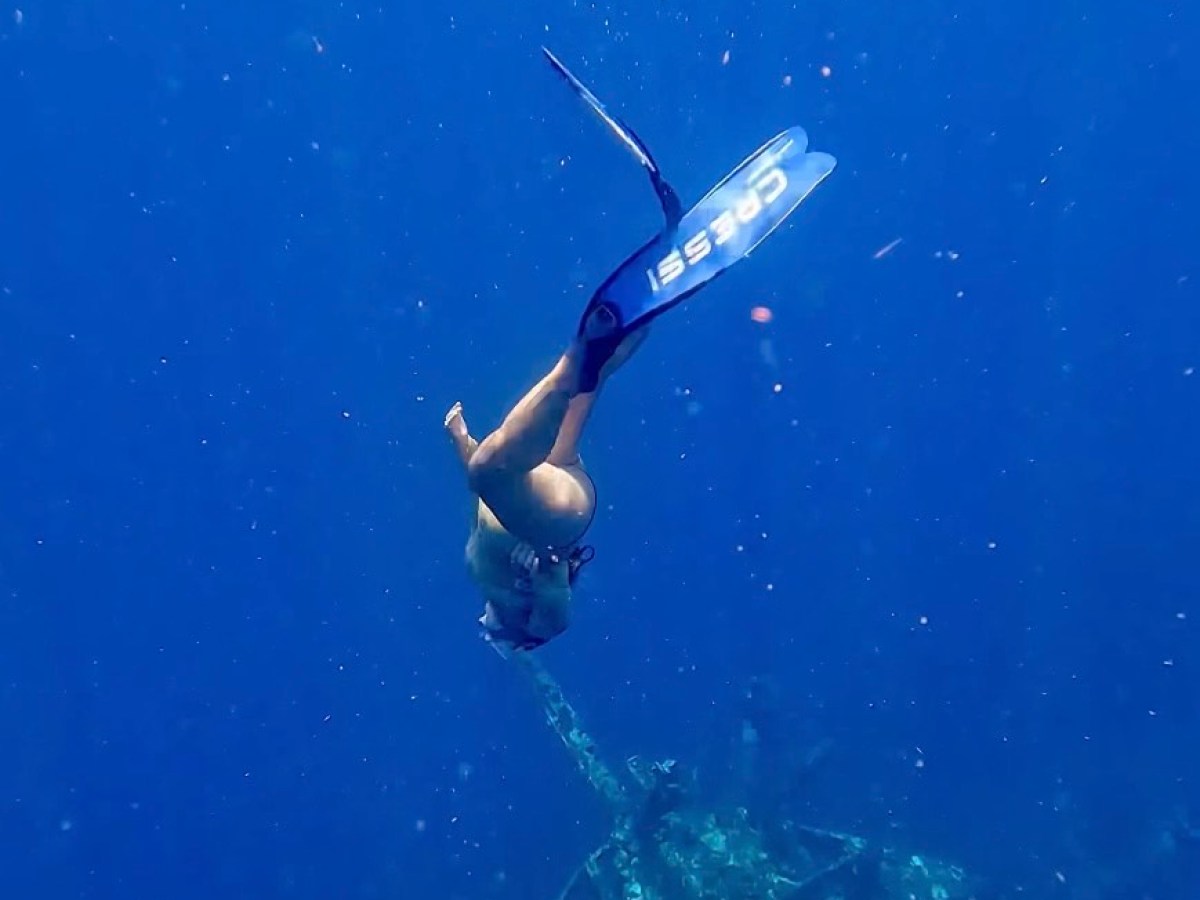 Diver with fins swims above underwater structure in deep blue sea.