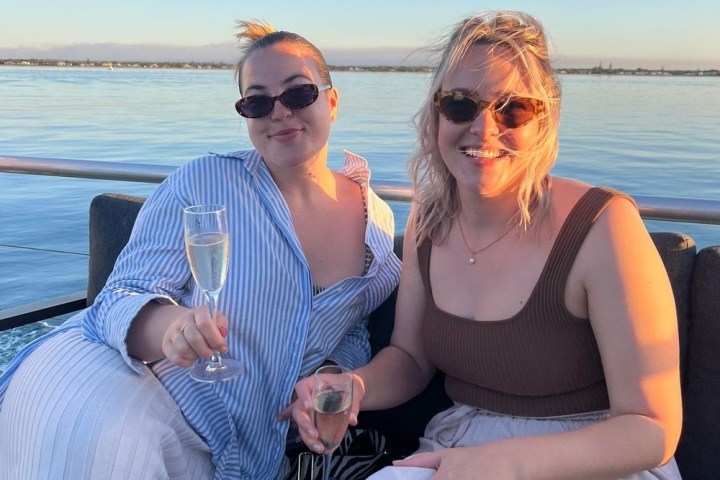 Two women in sunglasses on a boat, holding champagne glasses, with water and blue sky in the background.