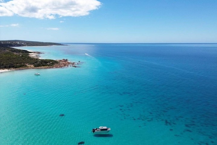 Aerial view of a boat on clear turquoise sea near a rocky coastline under a blue sky with few clouds.
